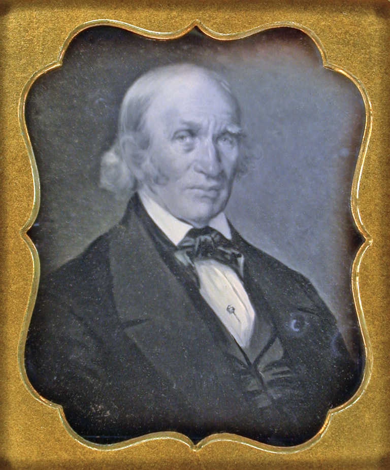 A black and white photograph of a white man in a high-collared shirt, necktie, vest, and jacket. The photograph is centered in a gold frame with inwardly curved edges.