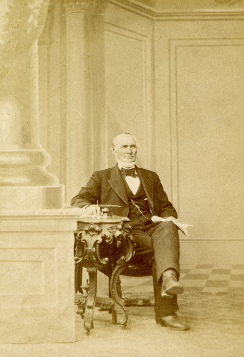 A black and white photograph of a man in a high-collared shirt, necktie, vest, jacket, pants and boots with his legs crossed. His left arm rests on a table with curved and ornately decorated legs. His right arm rests on his lap, and he is holding a bunch of folded papers.