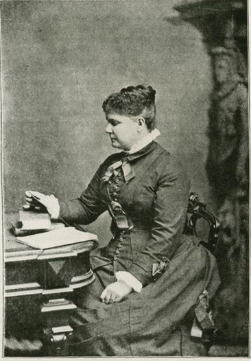 A black and white photograph of a white woman in a long, dark-colored dress. She looks at a document while sitting sideways, towards the left, at a desk.