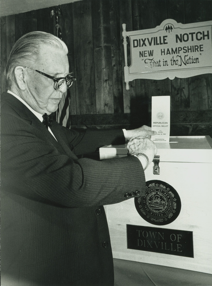 In this photograph, an older man with glasses and a suit and tie puts a ballot into a ballot box. The box with town seal reads "TOWN OF / DIXVILLE." Above the box in the black and white photograph is a sign reading "DIXVILLE NOTCH / NEW HAMPSHIRE / 