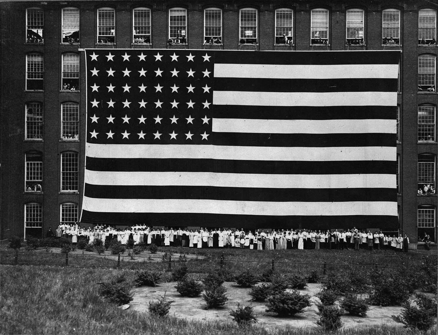 A black and white photograph of an American flag hanging against a multi-storied building. Groups of people can be seen hanging out the windows of the building, as well as at the base of the flag on the grass.
