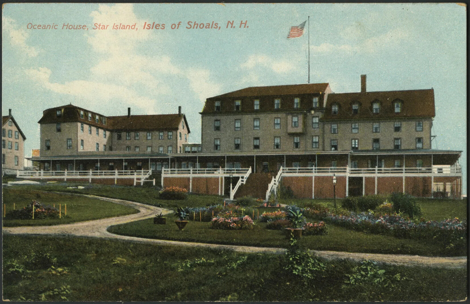 A colored postcard of three groups of buildings, and their surrounding green landscape. Two of these groups of buildings contain a long covered front porch. The main building, to the right-hand side, has an American flag on top of a tall flag pole. Clusters of colorful flowers dot the landscape and road, which curves from the left-hand side of the postcard and continues off to the right-hand side.