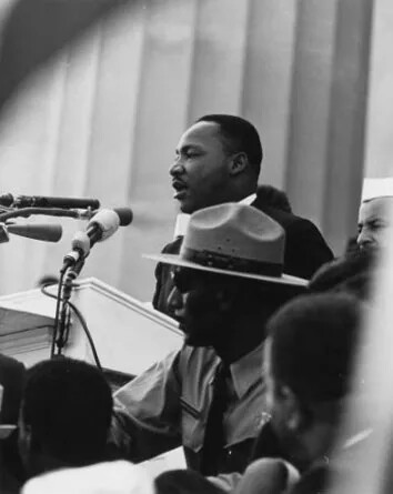 A black and white photograph shows a man wearing a suit speaking into several microphones. In front of him is an officer in uniform with large brimmed hat. There are other people listening, and the backdrop is a large column.