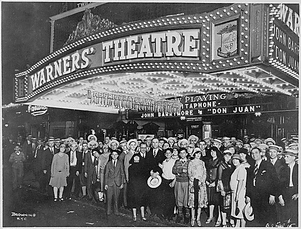 A black and white photograph of a large group of people standing in front of a movie theatre. They are all looking at the camera and smiling. The sign above the movie theatre is all lit up with small light bulbs.