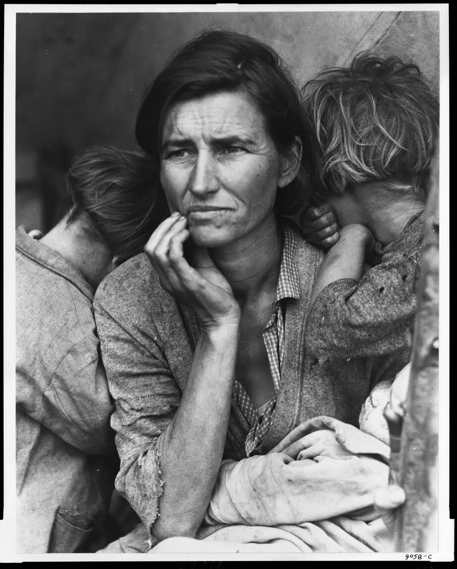 In a black and white photograph, a woman stares off into the distance, looking worried or tired. Her upper torso, shoulders, and head are seen, and her hand is holding her chin. Her hair is loosely pulled back, and she wears a blouse with cardigan. A small child is on either side of her with mussed hair and worn clothing. They have their heads on her shoulders and are facing away from the camera.