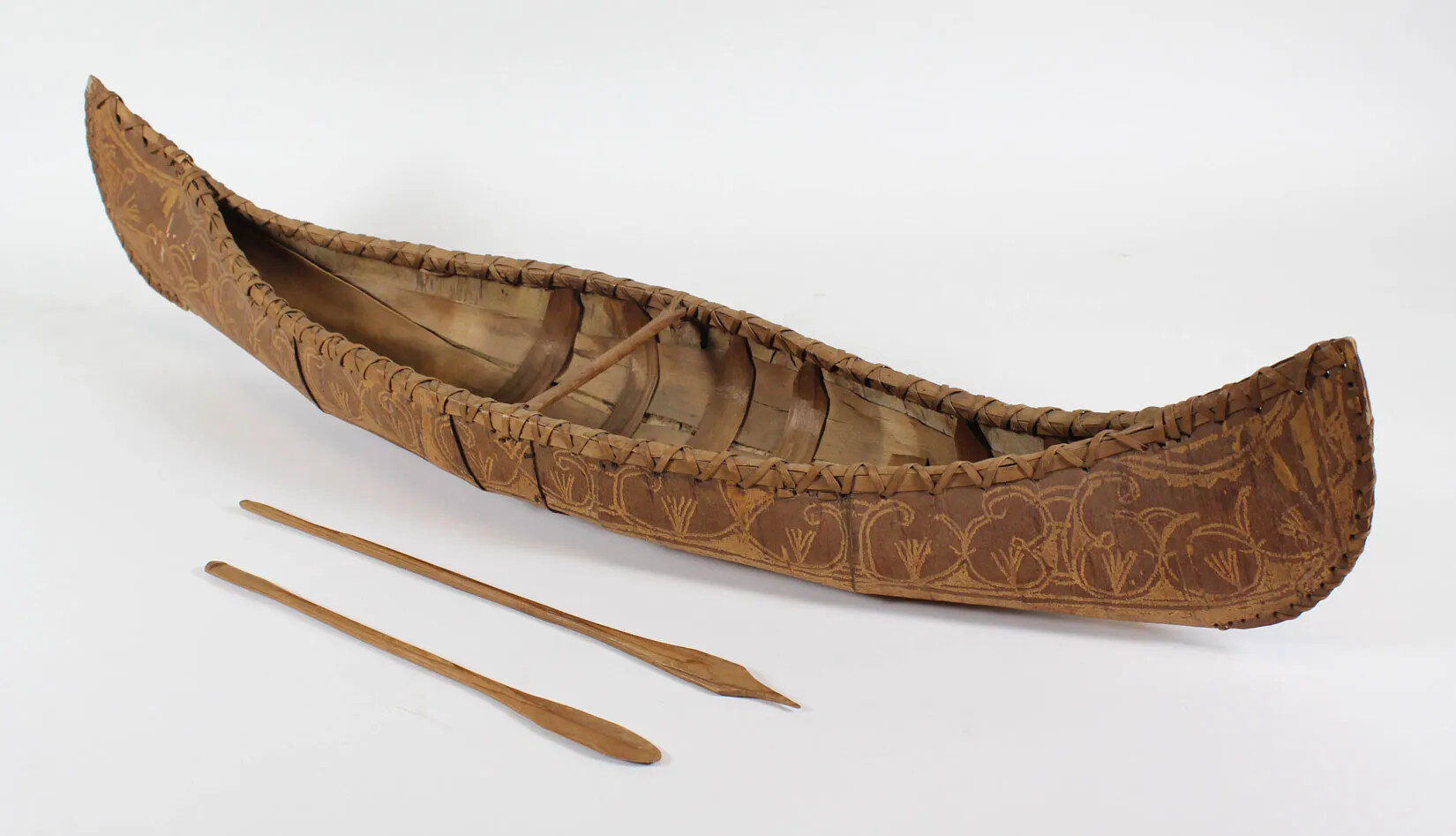 A model of a birchbark canoe has brown birchbark on a wooden frame. There is one thwart in the center. Intricate double-curve designs are on the outside of the canoe. Two wooden paddles lie beside the canoe, one with a rounded blade and one with a pointed blade.