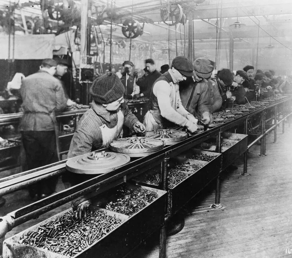 In a black and white photograph, many people work in an assembly line. They are in some sort of factory, and on their line are circular metal pieces, about 18" in diameter. Under the line are boxes of small pieces of metal, and they reach into the boxes to attach pieces to the circular metal. The workers wear hats, and long sleeve shirts or jackets. Some wear aprons. All are looking down at the assembly line