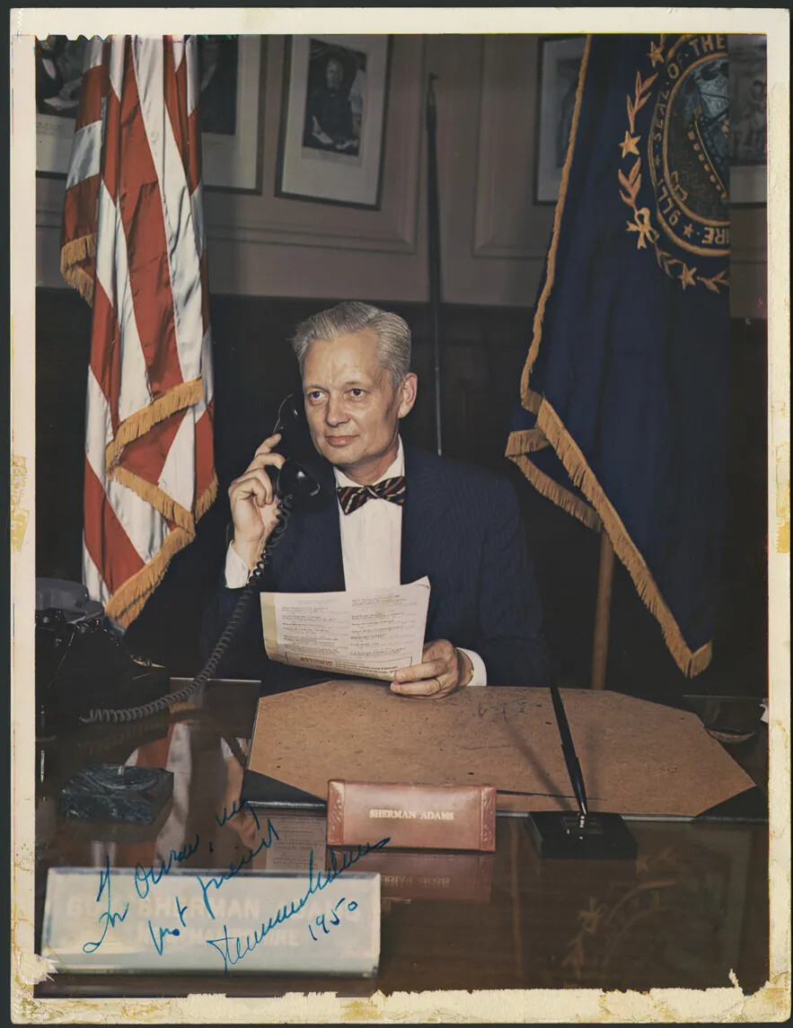 A white middle-aged man sits at a wooden desk with a corded phone in one hand and papers in the other in this color photograph. He looks off to the side. A United States flag and a New Hampshire state flag are standing on either side of him.