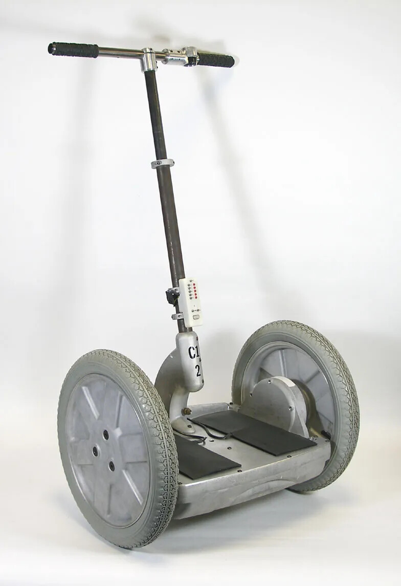 An aluminum deck, 17" wide x 15" deep, has two black rubber foot pads. It is supported by aluminum wheels with gray rubber tires, 16" in diameter. An adjustable steel column supports steel handlebars that have two black rubber handle grips. There is a control knob on the right handlebar and a white plastic control box at the bottom of column, with switches and red lights. A red nylon cord runs from a red "kill switch" at the bottom of the column to a black nylon strap on handlebar.