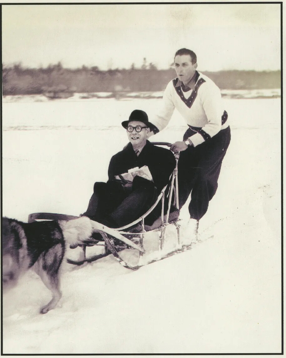 A photograph of two men, one seated, are on a dog sled in the wintertime being pulled by at least two dogs.