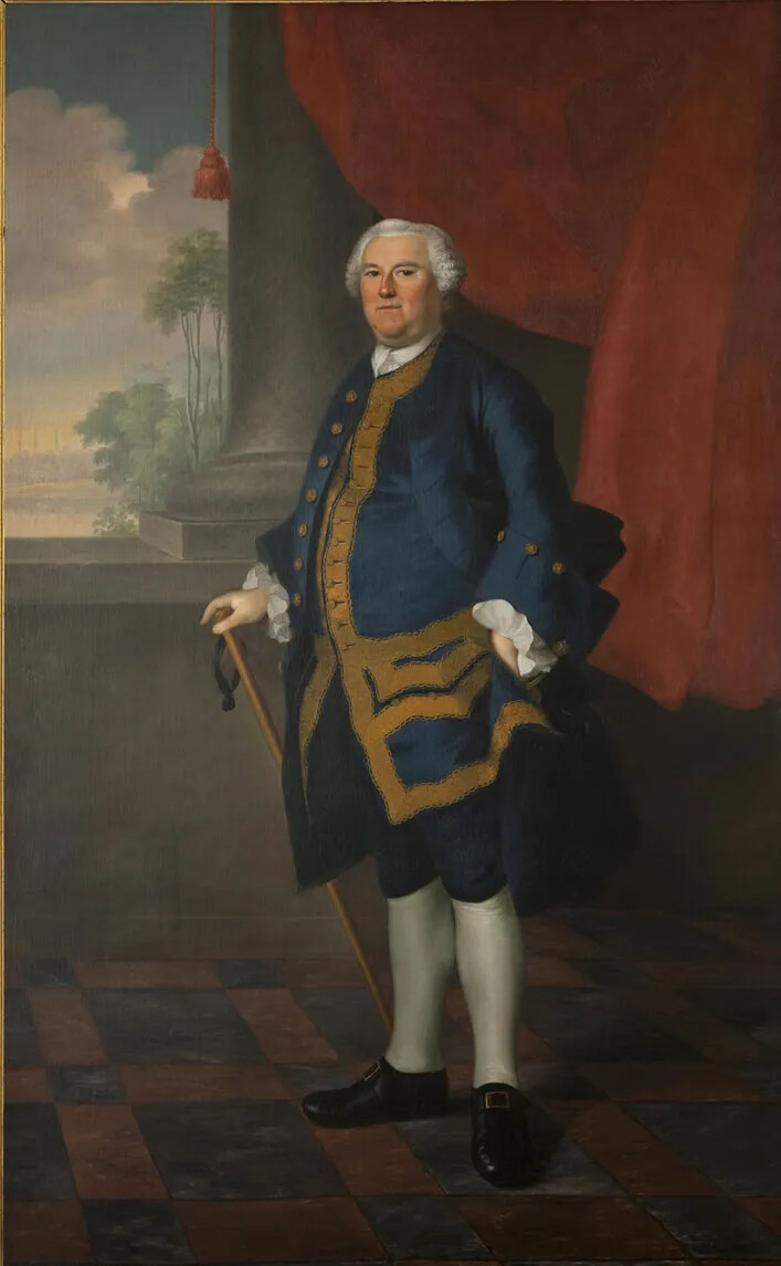 A colorful painting shows a white-haired man in formal dress posed standing with a cane. He is not smiling and looks directly at the viewer. He is wearing a long blue coat bordered with gold, a white shirt with ruffled cuffs, and knee-length blue pants. He has white stockings and black shoes with brass buckles. He stands on a black and tan checked floor in front of a column and red curtain. In the background are tall trees, clouds, and a village far away.