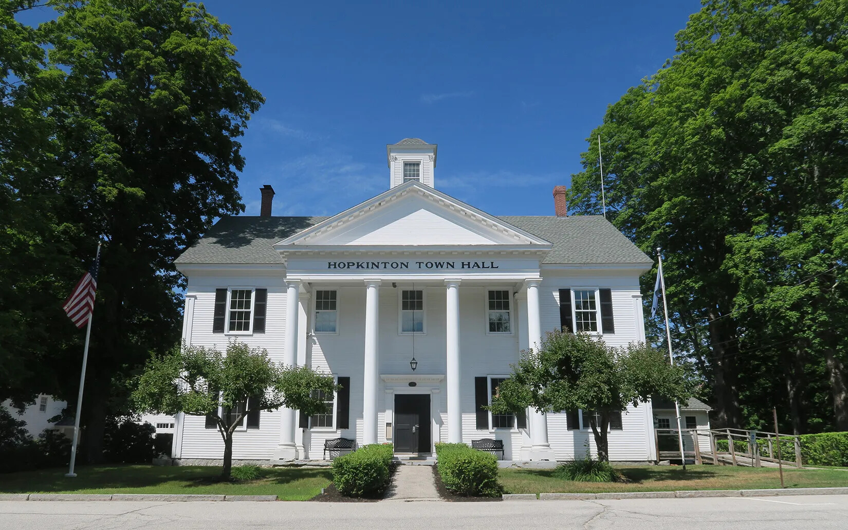 A two-story white building with black shutters. White columns in the front. Green shrubs line the walkway to the door. A sign above the door reads Hopkinton/Town/Hall in black letters. There is an American flag on a flagpole to the left of the building.