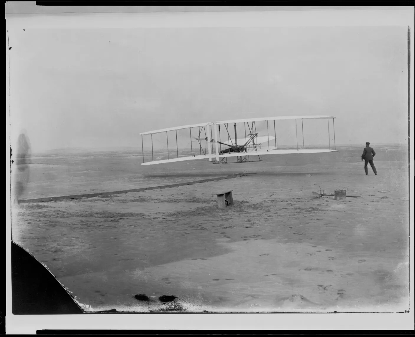In a black and white photograph, a biplane is shown with two wings stacked on each other. A small engine is seen along with a small body of the plane behind the wings. The plane is on a beach and a man stands watching it, near a wooden bench.