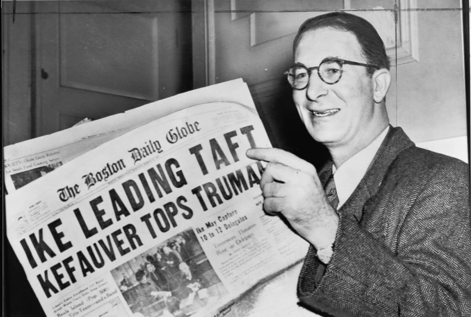 A black and white photograph of a white man smiling and holding an opened newspaper. The title of the newspaper reads "The Boston Daily Globe/IKE LEADING TAFT/KEFAUVER TOPS TRUMAN."