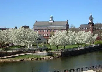 Historic Belknap Mill beside a river, with blooming trees and clear blue sky.