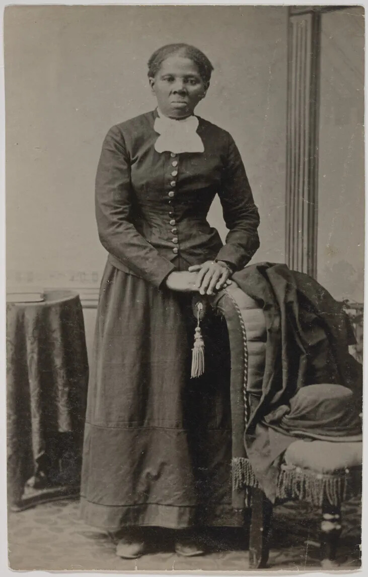 A formal black and white photograph shows a woman in dark mid-1800s clothing. Her hair is pulled back and she is not smiling. She is standing in a room between a brocade chair and a small, cloth covered table. She rests her hands on the back of the chair next to her. A jacket and woman's bonnet is on the chair.