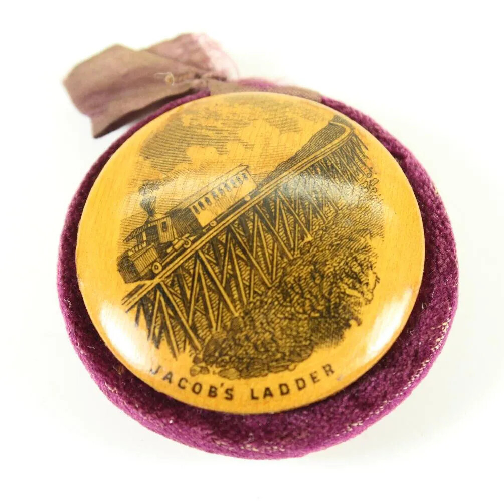 A wooden engraving of a train engine and one passenger car going up a very steep set of tracks on a mountain. The engraving is placed on a magenta colored piece of fabric, and has a faded pink ribbon in a loop at the top.