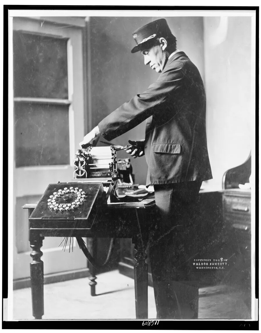 In this black and white photograph, a man stands in a room with a window. He wears a uniform with a cap decorated with gold braid. He stands at a desk, cutting paper from a machine on the desk. The machine has rollers and, on the side, a black square at waist height tilted up. The square is roughly a foot on all sides, and has two circles of buttons, one inside another.
