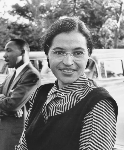 In a black and white photograph, a woman looks at the camera with a small smile. She has glasses, a striped blouse with a vest, and her hair pulled back. In the background, there is a car, and a man with a suit and tie, who is talking with his arms crossed.