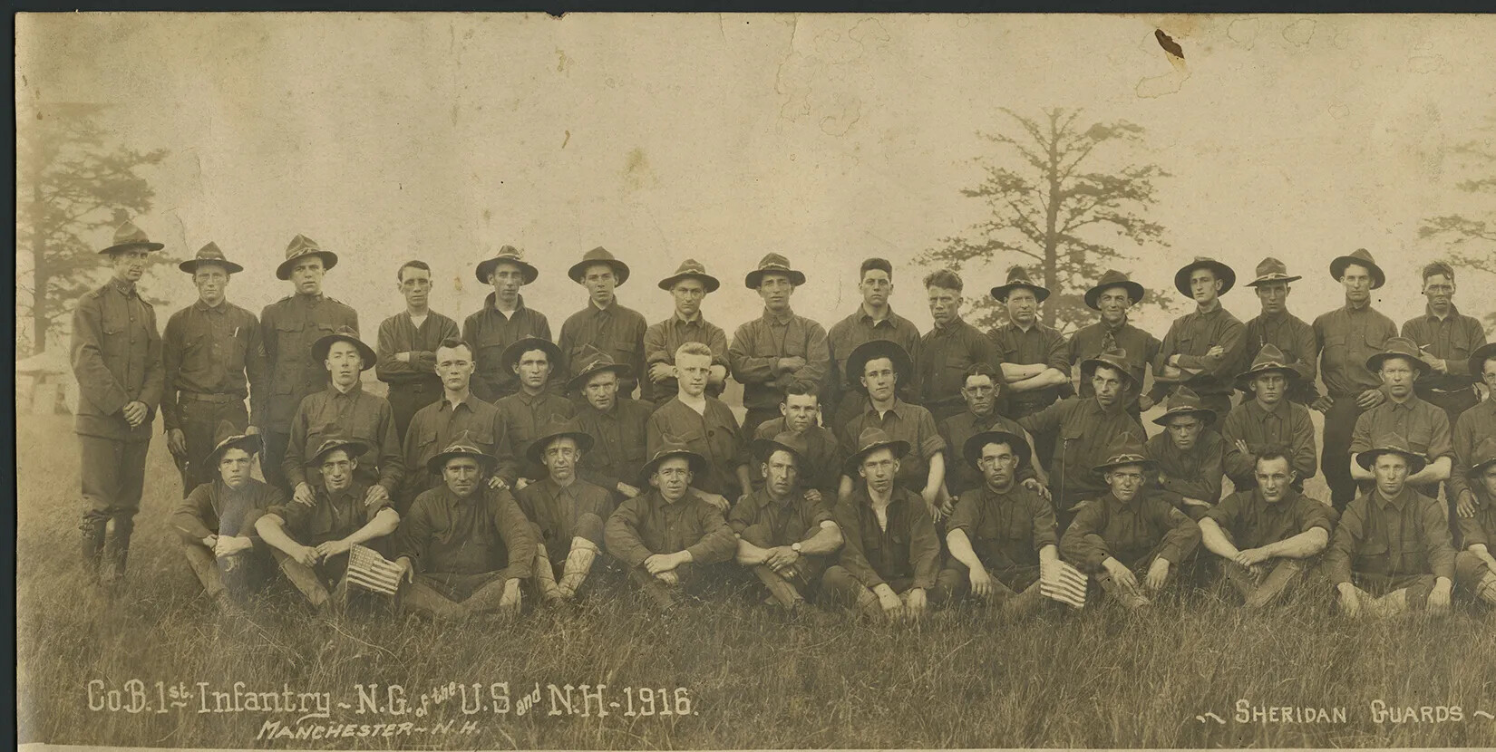 A black and white photograph of a group of men standing and sitting in three rows. They all wear military outfits and wide-rimmed hats. They are posed looking at the viewer in a grassy area. Tall trees can be seen in the background.