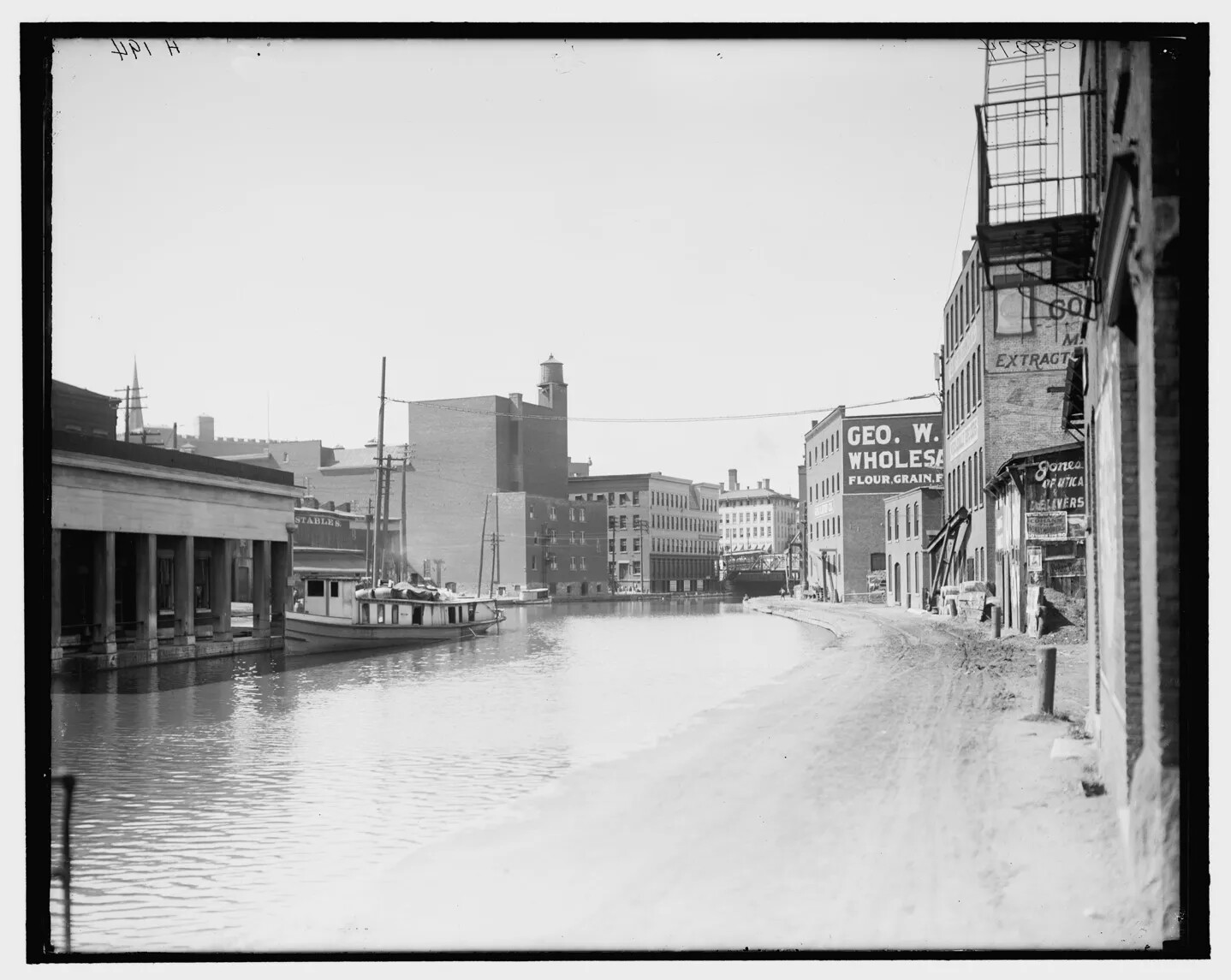A black and white photograph shows a wide, calm water canal curving through a developed town. On the right side, the water is bordered by sand and then buildings. On the left side, the water rises directly up to buildings. There is a boat tied up to one building.