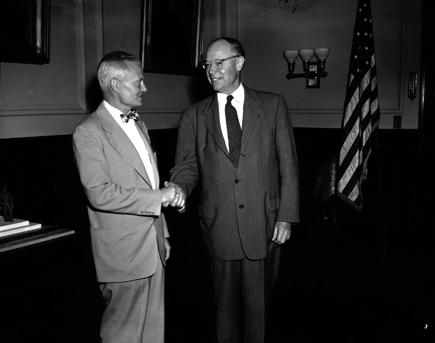 In a photograph, two white men in suits shake hands and smile.