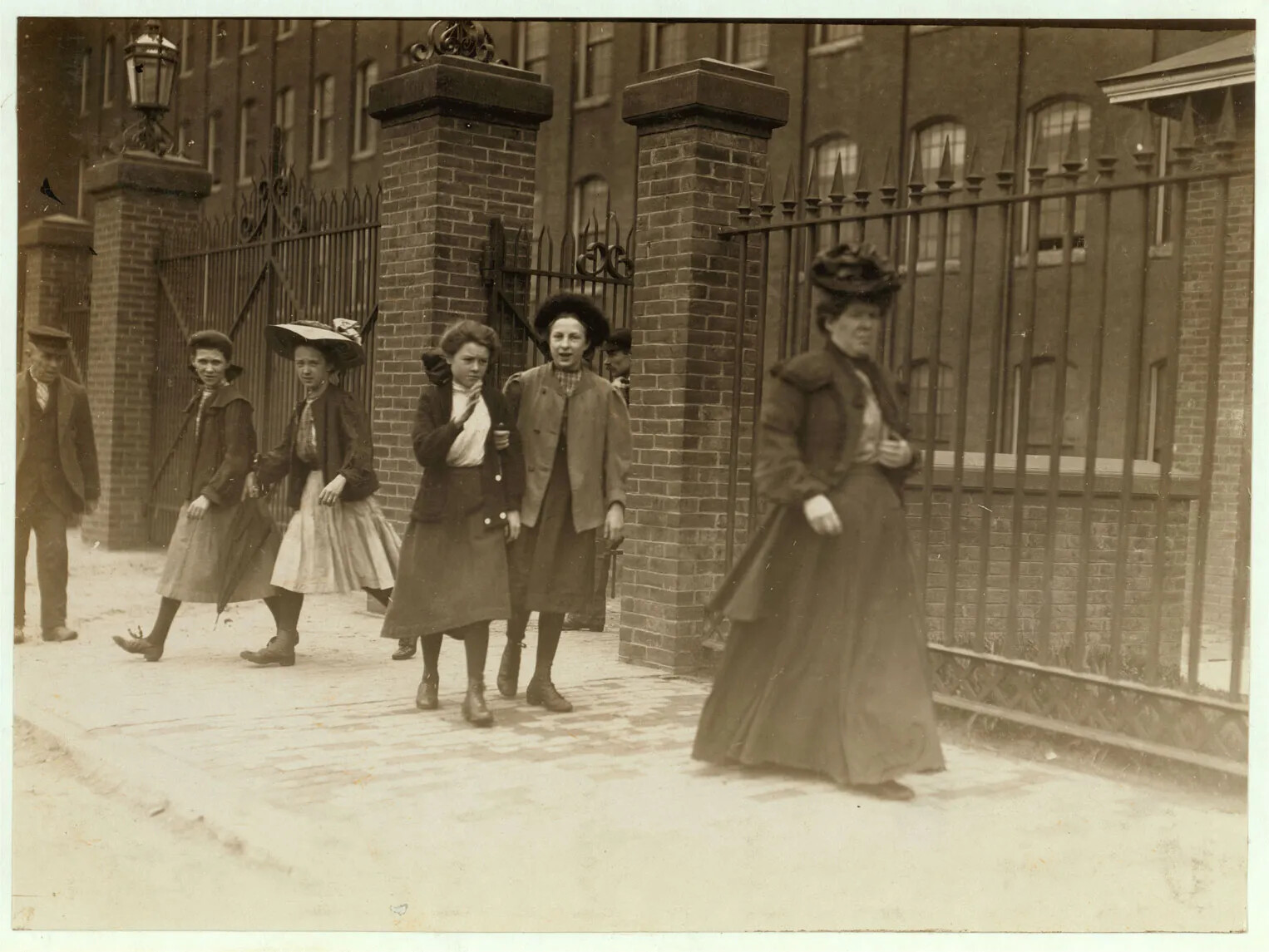 In a black and white photograph, the outside of a brick building is shown. Wrought-iron gates with tall brick columns fence the building. People are exiting through the gate out to the dusty sidewalk. One women wears long dark skirts, a formal jacket, and a decorative hat. Behind her, four girls just leaving the gate wear shin length skirts, jackets, and hats or bows in their hair. One man wears a dark suit and a casual hat and is watching them all leave.