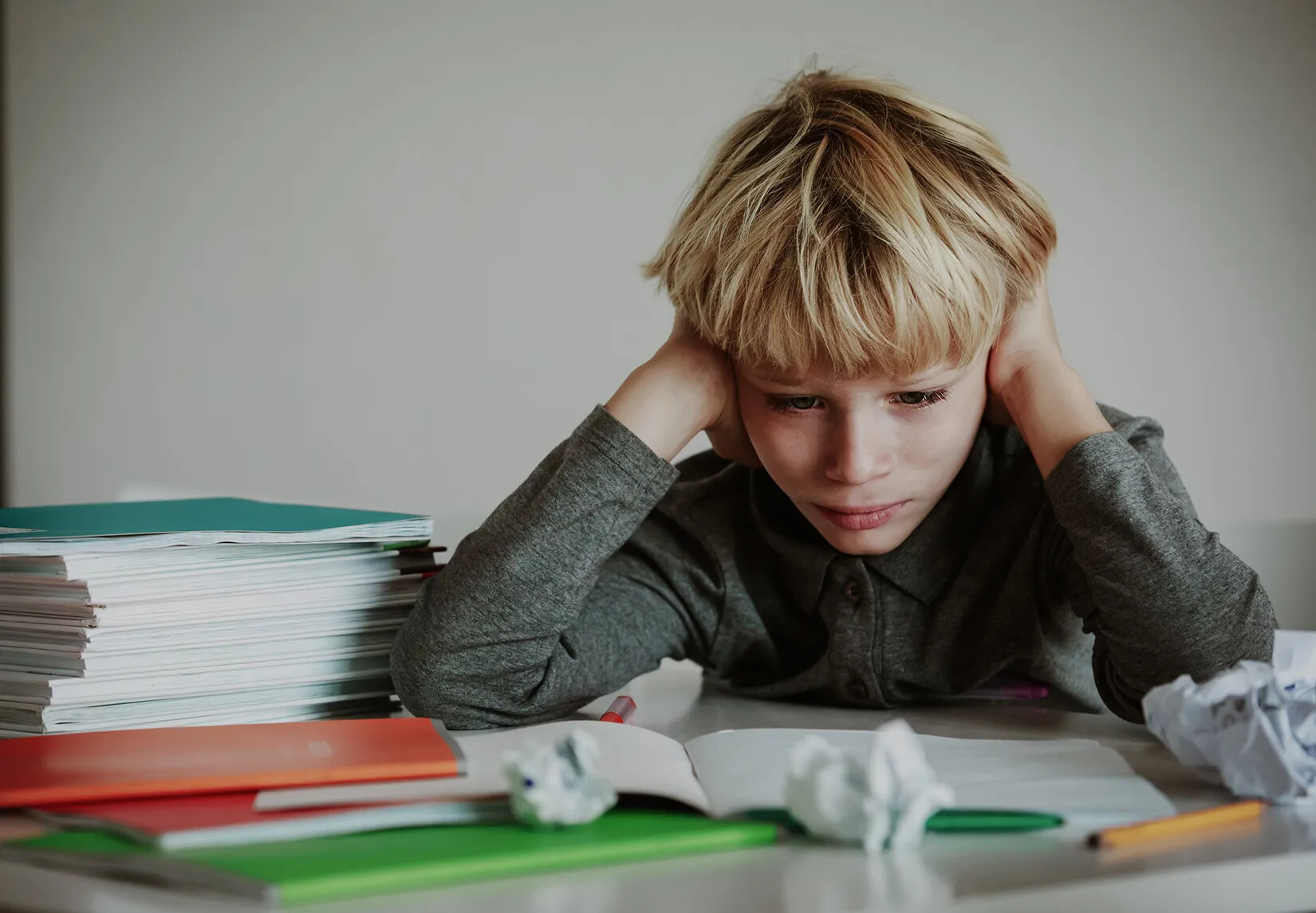 A colored photograph of a seated young white boy with his head in his hands, looking down at an open workbook. The table at which the boy sits has a pile of books to his left, and several notebooks and crumpled pieces of paper spread out in front of him.
