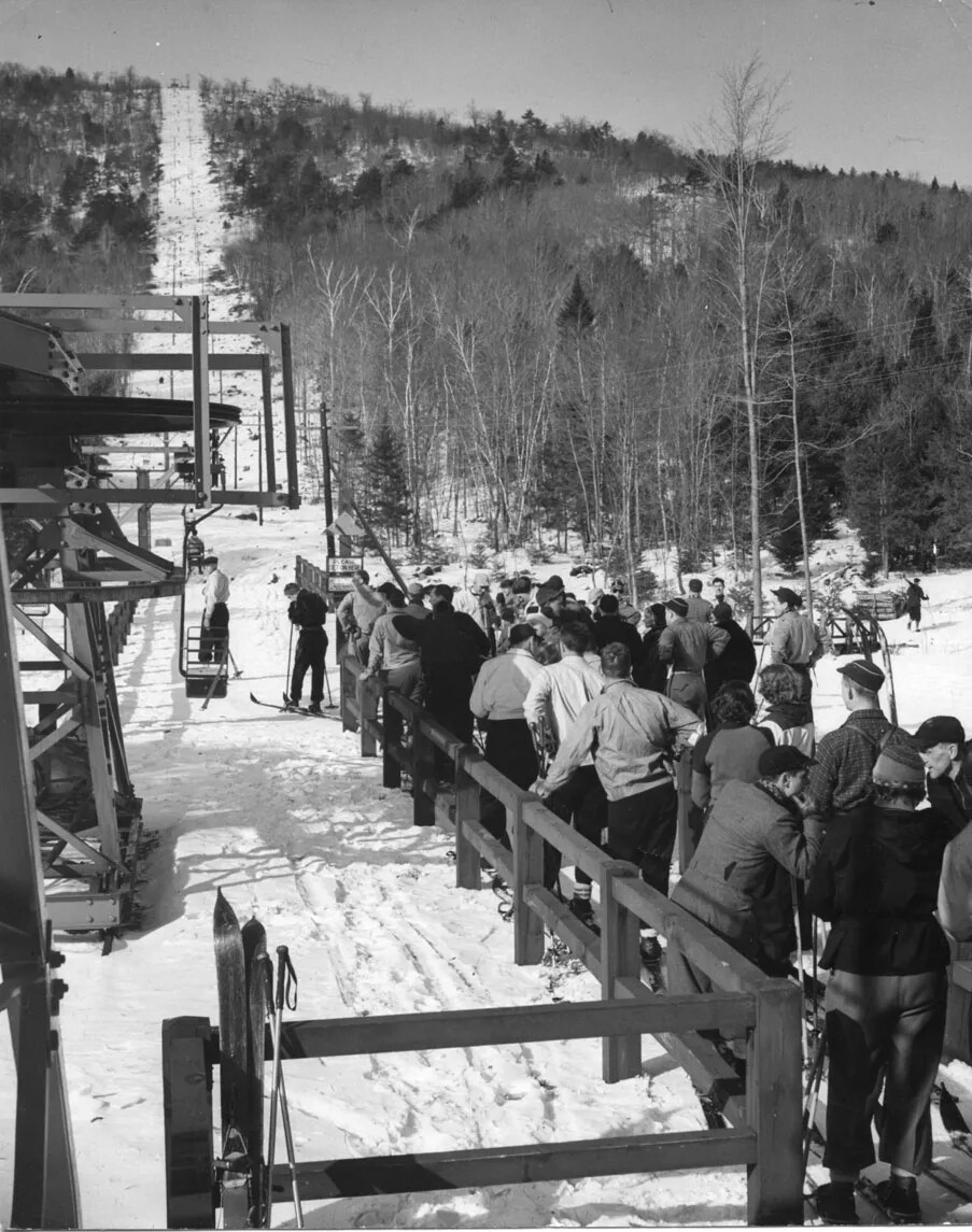 A black and white photograph shows a small mountain in the winter. Up the mountain runs a wide, white trail as well as supporting posts and wires for an old chair lift. In the foreground, people in winter clothes line up along a fence. The viewer can see one person getting ready to board the next chair that is coming.