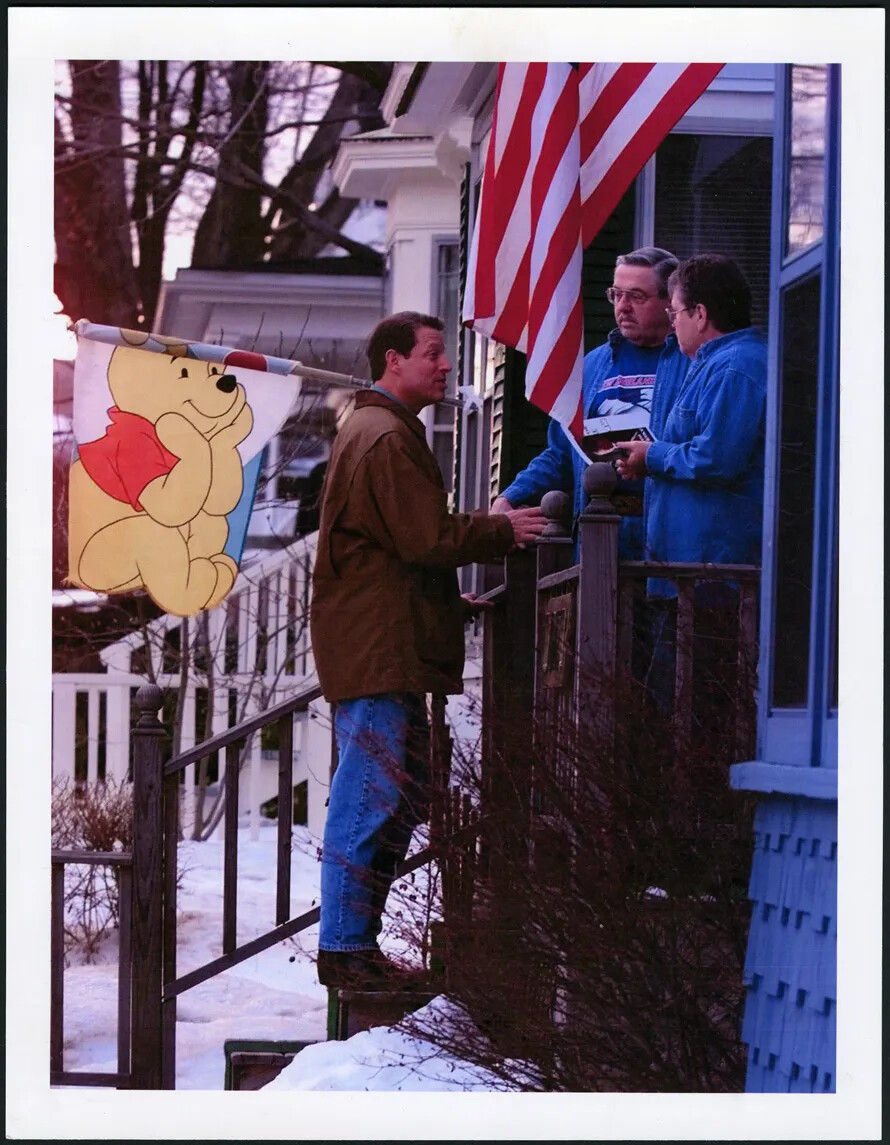 In a photograph a man in a jacket and jeans stands on steps leading up to the front porch of a house. He speaks to a man and woman, and she holds a pamphlet with a picture of him on it. Part of an American flag is visible hanging just next to the group. Snow covers the ground and the bushes and trees are bare.