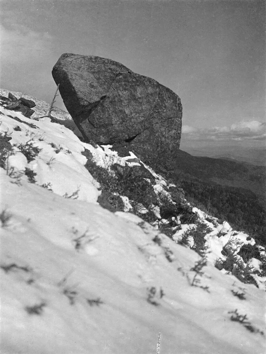 On a snow-covered steep hill a large, irregularly-shaped boulder sits perched in this photograph.