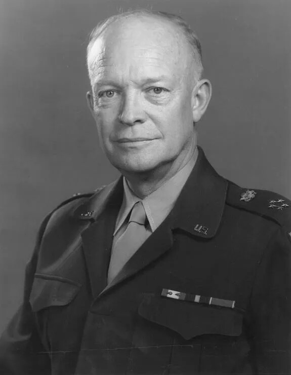 A black and white photograph of a white man, looking at the camera. He wears a collared shirt and tie, and a military-style jacket.