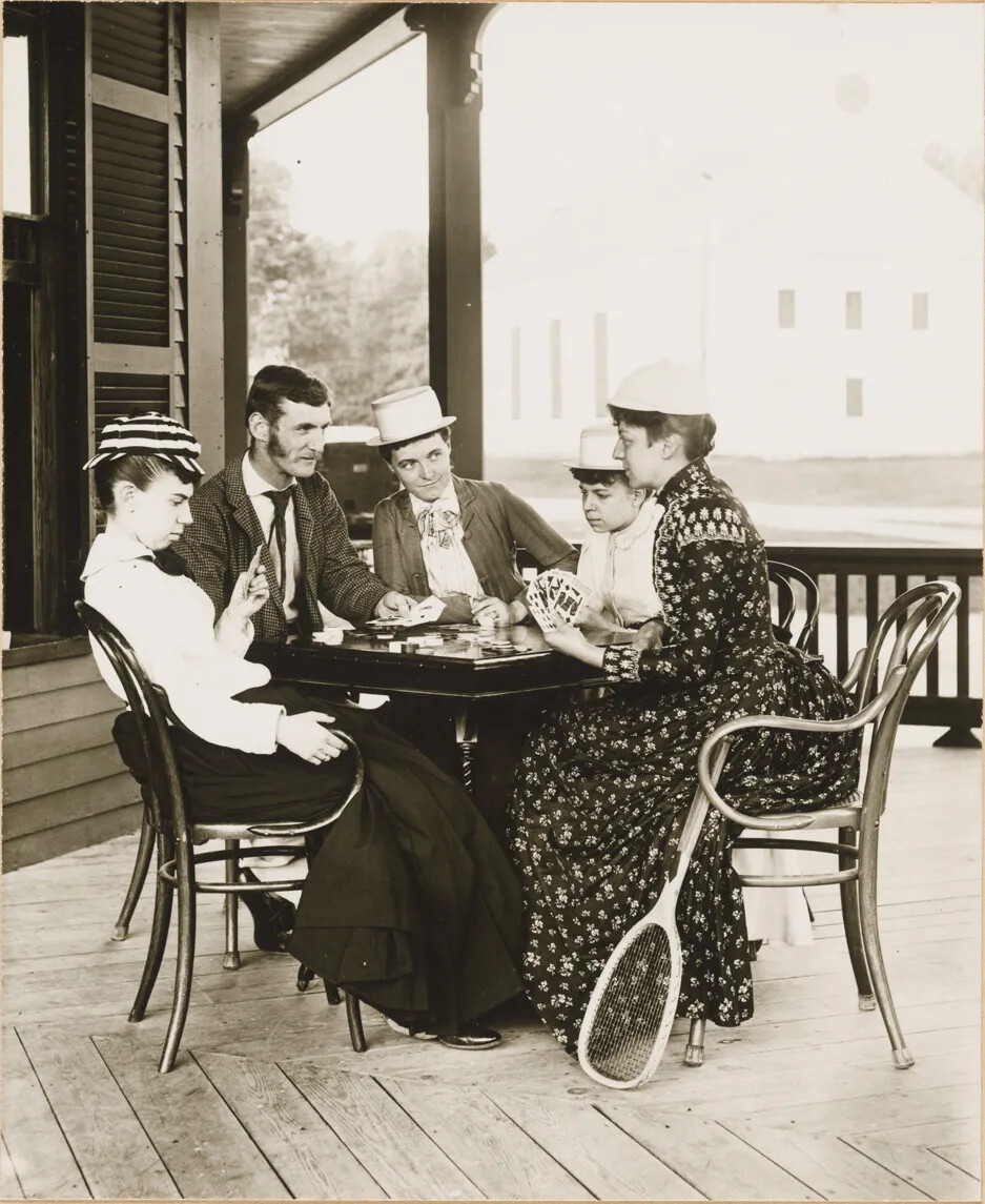 Black and white photograph of four women and one man sitting around a table playing cards on a large porch.The woman seated at the photograph's right-most corner has a tennis racket resting against her chair.