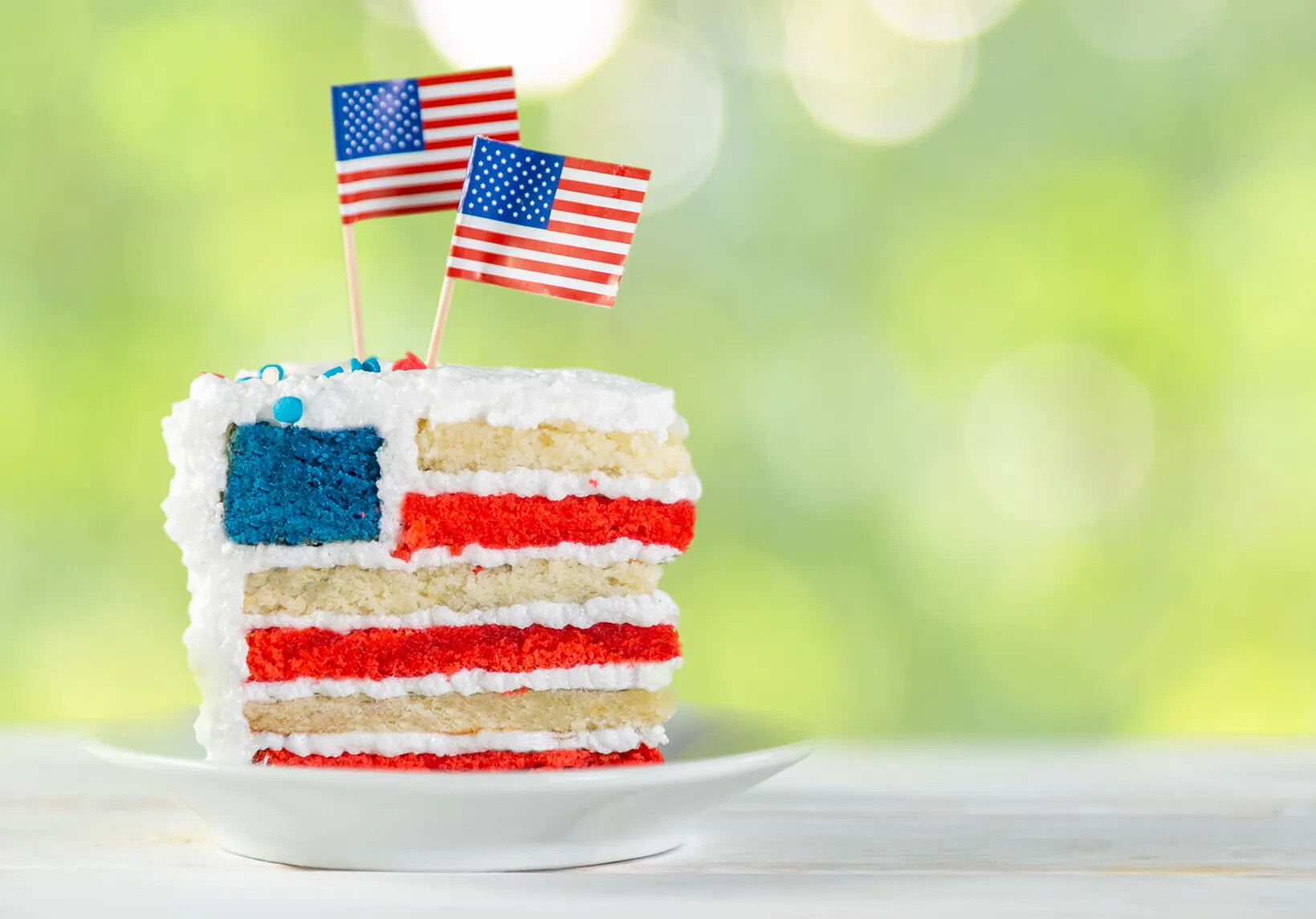 A colored photograph of a piece of cake that is designed to look like the American flag, with strips of red, white, and blue cake. Sticking out of the top of the cake are two American flags attached to toothpicks.