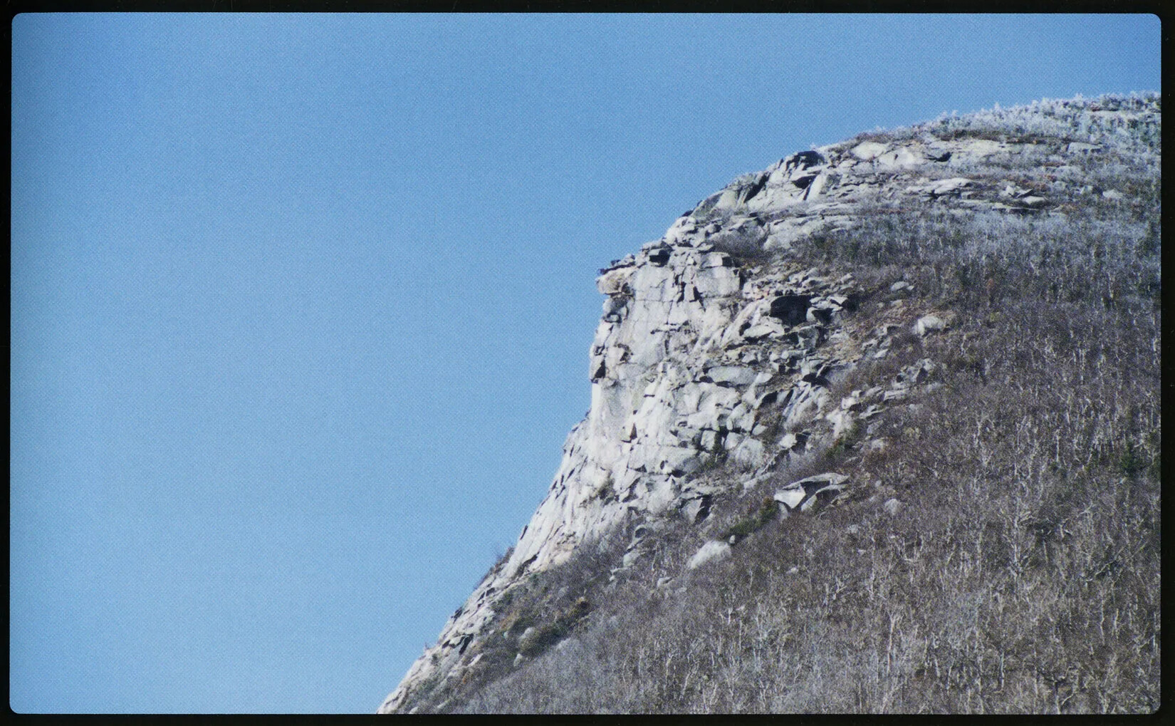 The edge of a rocky mountain is seen in this color photograph. There are sparse trees leading to the rocky cliff, which has granite slabs of no particular formation.