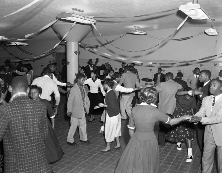 A black and white photograph shows a large room with a pillar and some decorations. Teenangers dance in the room in pairs. Boys wears jackets and ties and girls wears skirts or dresses.