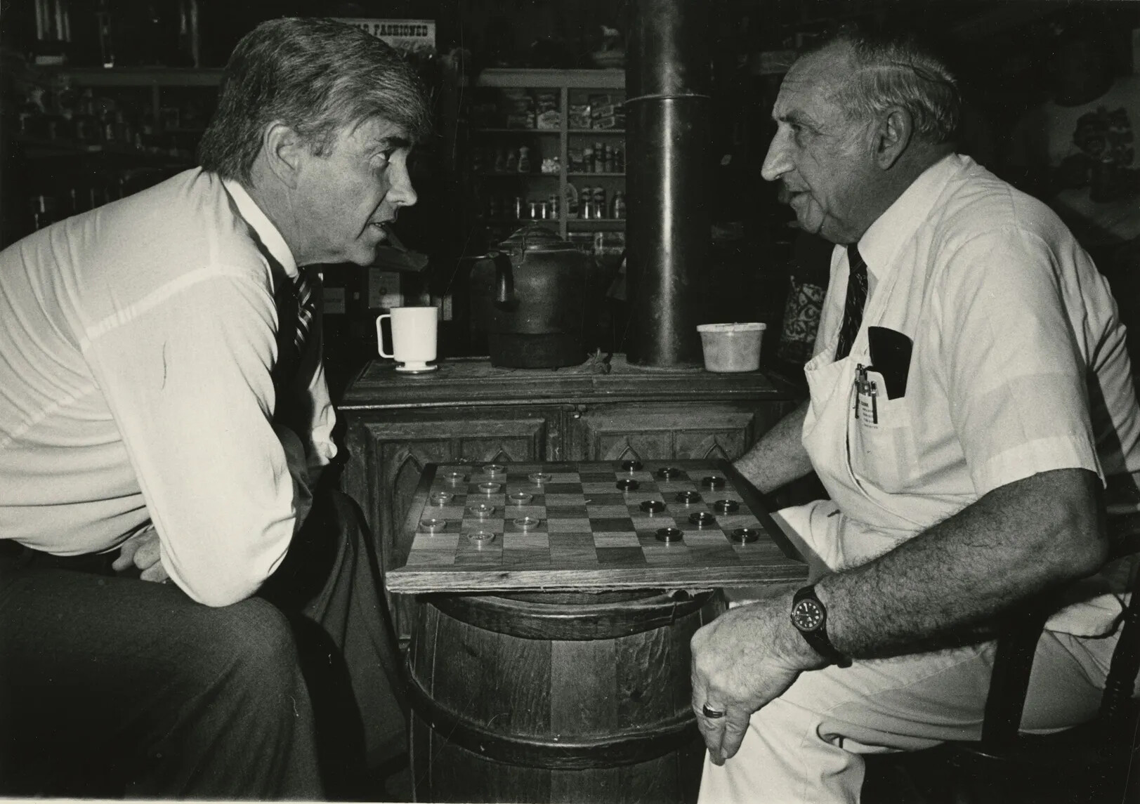 In a photograph two men sit facing each other over a checkerboard. The man on the left is wearing a buttondown shirt and tie. The man on the right has a short sleeve buttondown shirt, tie and a white apron.The checkerboard sits on a barrel in front of a wood-burning stove. Shelves with canned and dry goods appear in the background.