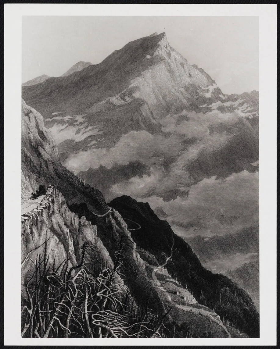 A black and white photograph shows steep rocks rising to the left in the foreground. In the distance, a large mountain is seen, rising through clouds. The peak is rocky and bare.