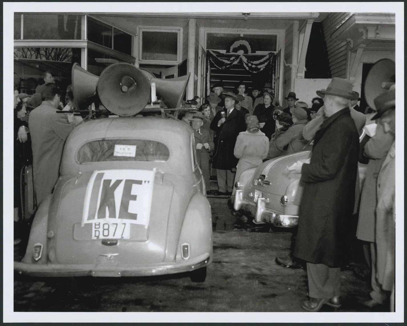 In a photograph of a street scene a man in an overcoat and hat stands near an open doorway on a sidewalk, holding a microphone and speaking to a crowd of people gathered around him. People wear raincoats, women are wearing hats or scarves over their heads, and men are wearing hats. A car parked at the curb has loudspeakers on its roof and a sign reading "Ike" taped to the trunk.