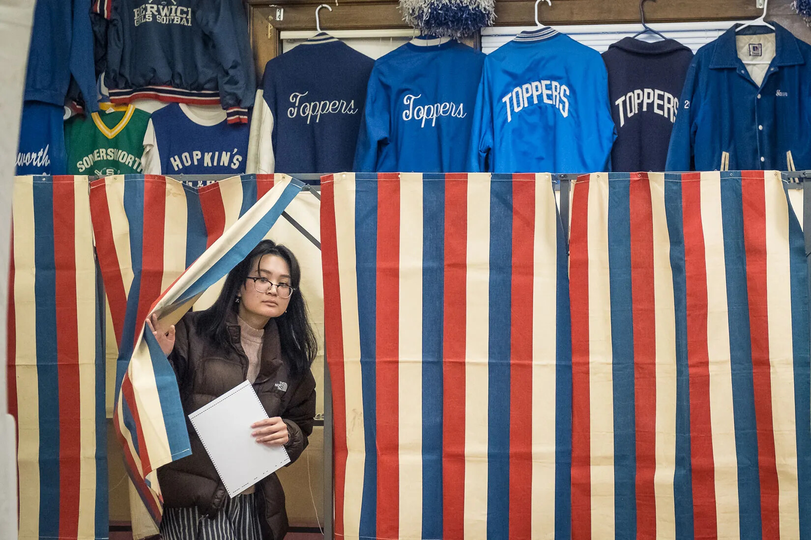 A woman steps out from a voting booth holding a piece of paper.  Blue sports jackets line the upper third of the photograph.