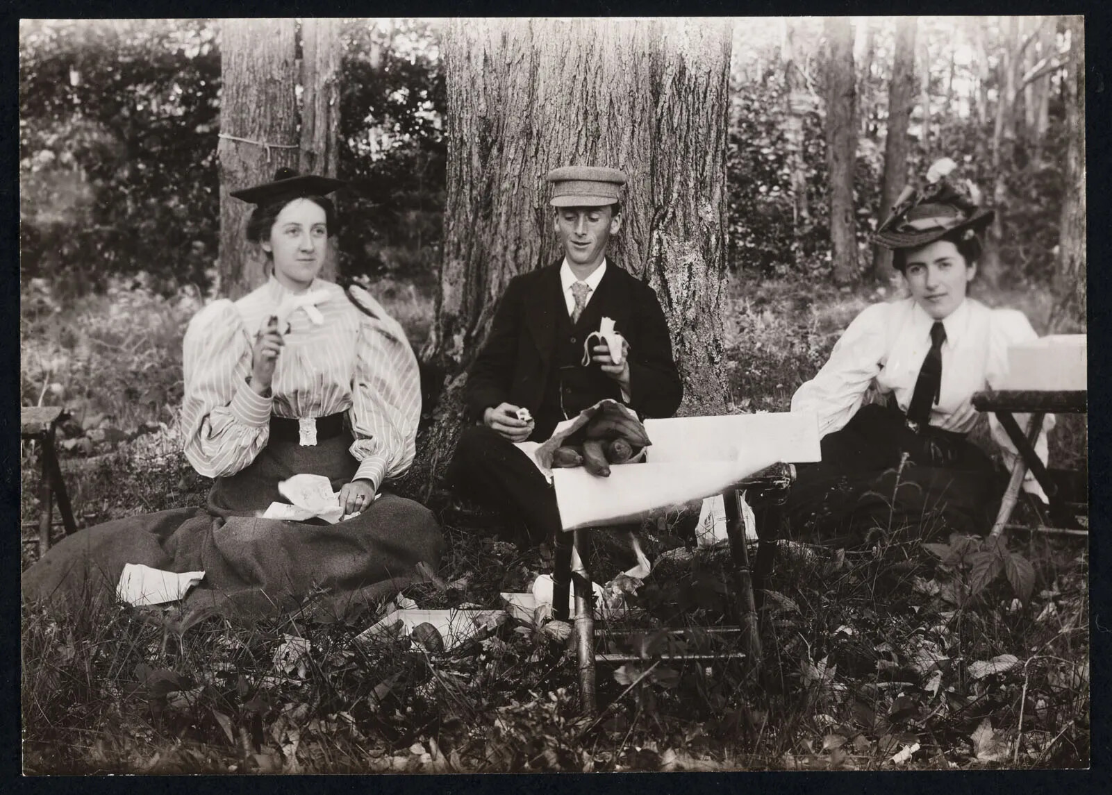 A black and white photograph of two women and a man sitting eating a picnic in the woods. There is a low table placed in front of the group, and on the table is a bag of food. The woman on the left and the man eat bananas. The two women wear shirts with puffy sleeves and long skirts and hats.