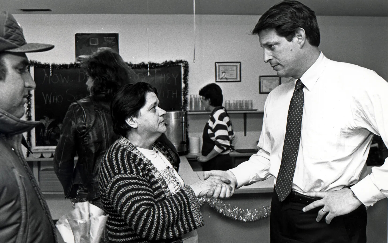 A man wearing a buttondown shirt and tie stands with hand on hip while shaking hands with a shorter woman. Woman wears sweater and t-shirt, and other man stands to her left in profile. Glasses and silverware containers are seen in the background of this photograph.