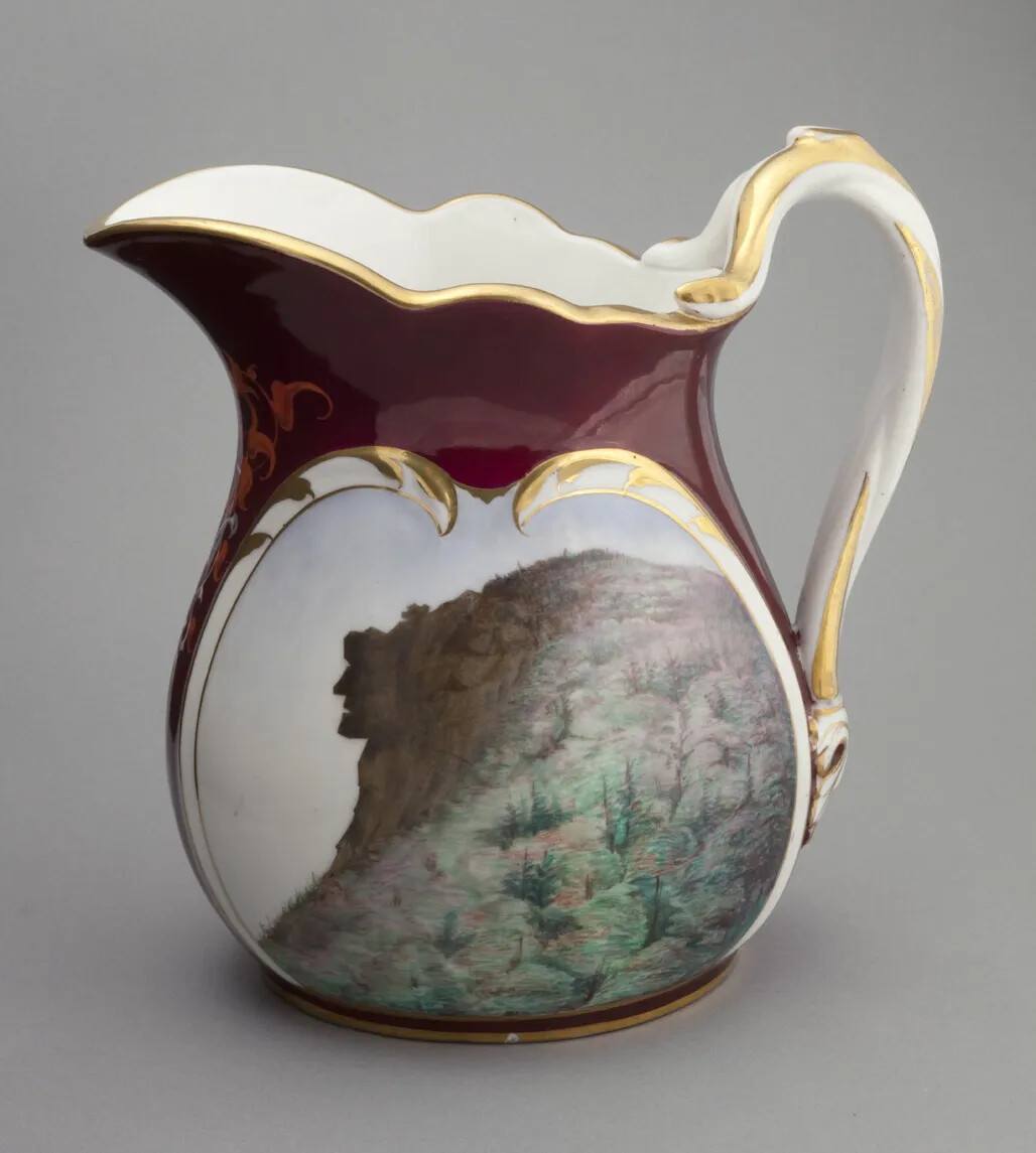Molded, white glazed porcelain pitcher has a tapered neck, wide mouth and spout, and loop handle. On the body of the pitcher is a printed image of a painting of a profile of a man's face on a mountain top. The picture is in an oval floral medallion panel. The pitcher has a maroon background with decorative red, white, and gold mirrored scrolls.
