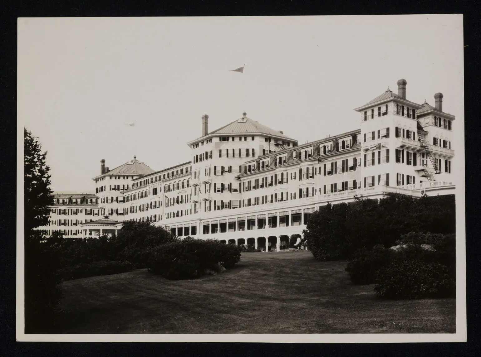 A black and white photograph of a large building. The building hasa long covered porch, and several circular and rectangle towers. In front of this building is a grassy area and several bushes.