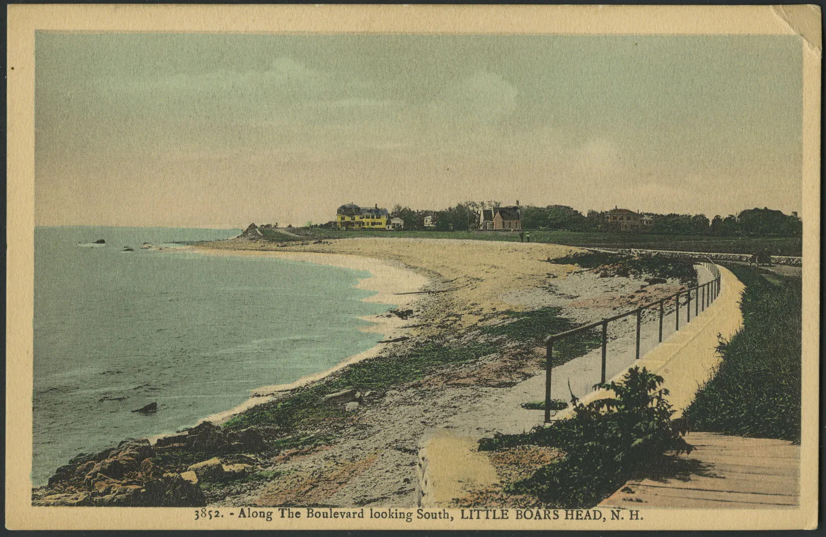 A colored postcard of the ocean and a curved sandy shore. Houses are seen in the background of the road's curve. The sand at the center and bottom forefront of the postcard is covered in seaweed. A large border and fence curves along with the road on the right-hand side of the postcard. A car and two people walking further ahead share the road.
