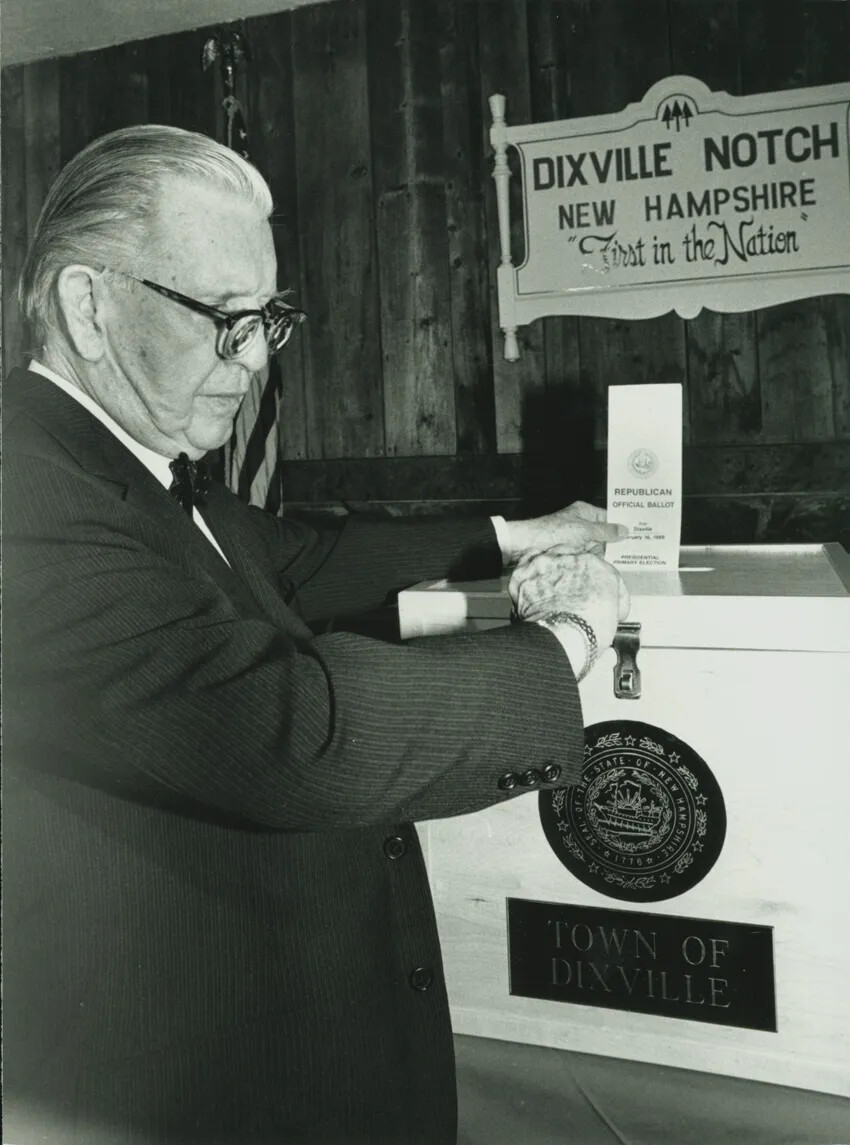 In this photograph, an older man with glasses and a suit and tie puts a ballot into a ballot box. The box with town seal reads "TOWN OF / DIXVILLE." Above the box in the black and white photograph is a sign reading "DIXVILLE NOTCH / NEW HAMPSHIRE / 'First in the Nation.'"